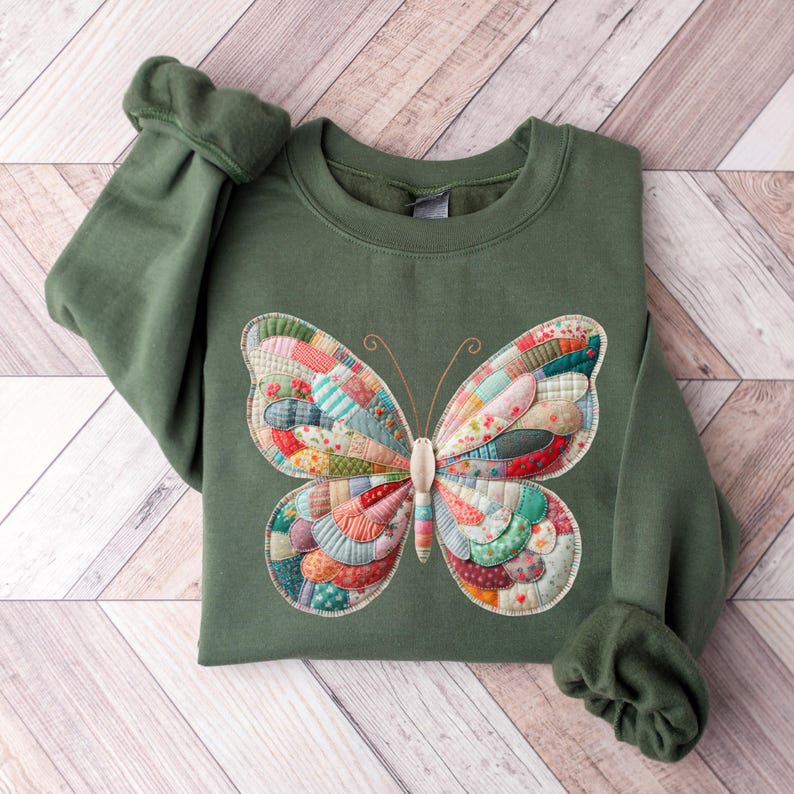 May include: A green sweatshirt featuring a large, colorful butterfly applique. The butterfly design is made of various fabric patches in shades of red, blue, green, and yellow. The sleeves are cuffed, and the sweatshirt is folded on a wooden surface.