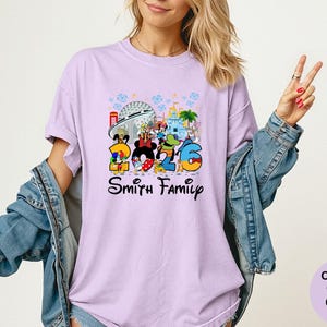 May include: A light purple t-shirt featuring a colorful graphic of Disney characters and the number "2026". The text "Smith Family" is below the graphic. The shirt is a Comfort Colors Orchid.