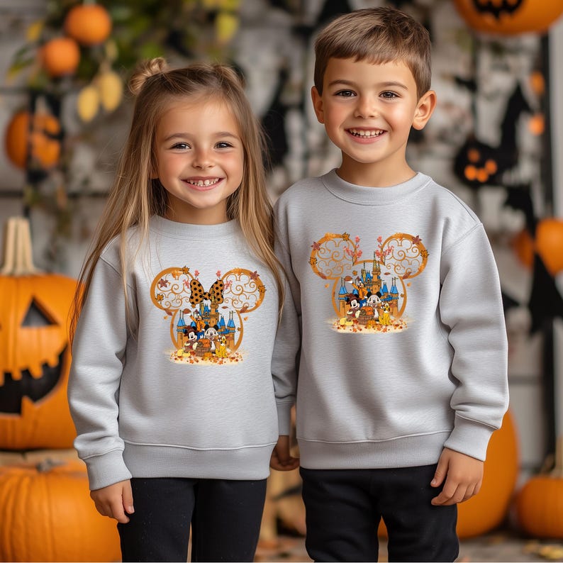 Pode incluir: Duas crian&ccedil;as vestindo moletons cinza claro com um design tem&aacute;tico do Mickey Mouse. O design apresenta um castelo, Mickey e Minnie Mouse e folhagens de outono. Os moletons s&atilde;o de manga comprida e adequados para o outono ou Halloween.