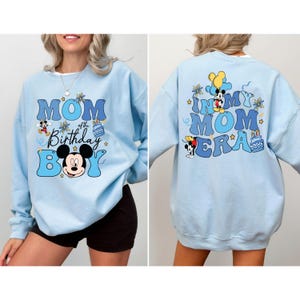 K&ouml;nnte beinhalten: Hellblauer Pullover mit einem grafischen Aufdruck von Mickey Mouse und dem Text "Mom of the Birthday Boy" auf der Vorderseite und "In My Mom Era" auf der R&uuml;ckseite.