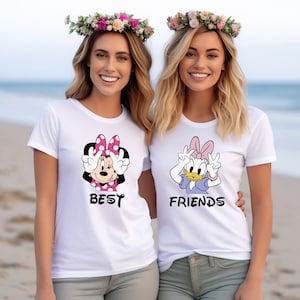 Może przedstawiać: Dwie białe koszulki z nadrukiem postaci z kresk&oacute;wek: Minnie Mouse i Daisy Duck. Koszulka z Minnie Mouse ma napis "Best", a koszulka z Daisy Duck ma napis "Friends".