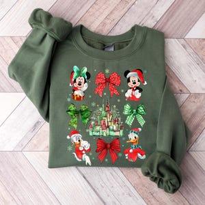 May include: Green Christmas sweatshirt with a festive design featuring Mickey Mouse, Minnie Mouse, Donald Duck, and Daisy Duck wearing Santa hats. The design also includes a red bow, a green bow, and a depiction of Cinderella's castle.