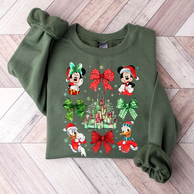 May include: Green Christmas sweatshirt with a festive design featuring Mickey Mouse, Minnie Mouse, Donald Duck, and Daisy Duck wearing Santa hats. The design also includes a red bow, a green bow, and a depiction of Cinderella's castle.