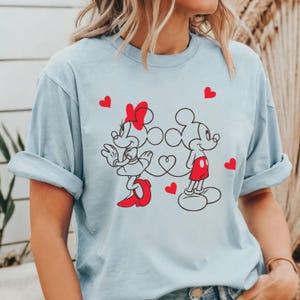 Op de afbeelding: Een lichtblauw T-shirt met een grafische print van Mickey Mouse en Minnie Mouse die hand in hand lopen. De grafiek is in rood en zwart en bevat harten.