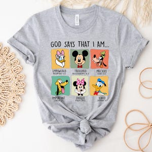 Puede incluir: Una camiseta gris claro con personajes de Disney y el texto "GOD SAYS THAT I AM...". La camiseta incluye im&aacute;genes de Daisy, Mickey Mouse, Goofy, Pluto, Minnie Mouse y el Pato Donald, con palabras y vers&iacute;culos b&iacute;blicos.