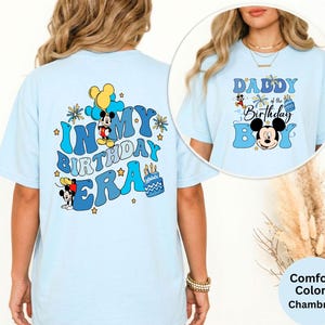 Op de afbeelding: Lichtblauw T-shirt met de tekst "IN MY BIRTHDAY ERA" en Mickey Mouse-afbeeldingen op de rug. De voorkant toont "DADDY of the Birthday BOY" en een Mickey Mouse-afbeelding. Het shirt is een comfort color chambray.