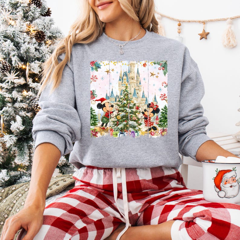 Może przedstawiać: Szara bluza z nadrukiem o tematyce świątecznej z Myszka Miki i Myszka Minnie przed zamkiem i choinką. Bluza jest połączona z czerwono-białymi spodniami od piżamy w kratę. Widoczny jest r&oacute;wnież kubek z motywem Świętego Mikołaja.