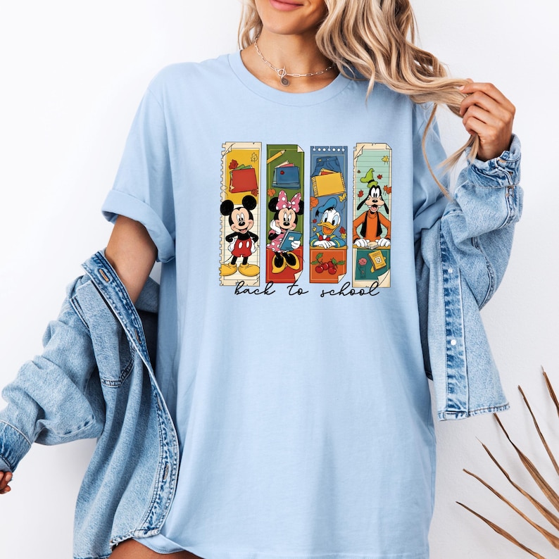 May include: Light blue t-shirt featuring Mickey Mouse, Minnie Mouse, Donald Duck, and Goofy in a back-to-school design. Each character is depicted in a colorful, vertical frame with school-themed elements. The words "back to school" are written in a script font.