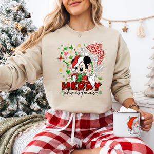 May include: Beige sweatshirt featuring a festive Mickey Mouse design with a Santa hat and scarf. The graphic includes the text "MERRY christmas" in a plaid pattern. The image also shows red and white plaid pajama pants.