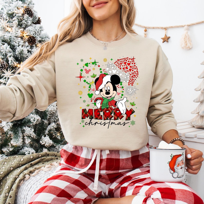 May include: Beige sweatshirt featuring a festive Mickey Mouse design with a Santa hat and scarf. The graphic includes the text "MERRY christmas" in a plaid pattern. The image also shows red and white plaid pajama pants.