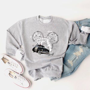 Puede incluir: Sudadera gris con una ilustraci&oacute;n en blanco y negro de un coche de polic&iacute;a vintage, orejas de Mickey Mouse y la palabra "Sheriff".