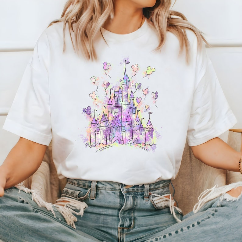 May include: A white t-shirt featuring a watercolour illustration of a castle in shades of purple, pink and blue. The castle is surrounded by balloons shaped like Mickey Mouse ears.