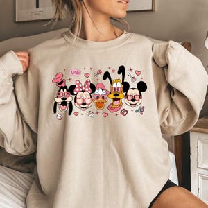 Op de afbeelding: Een beige sweatshirt met een grafische print van Mickey Mouse, Minnie Mouse, Donald Duck, Goofy en Pluto die roze hartvormige zonnebrillen dragen. De afbeelding bevat ook harten, sterren en het woord "Love!" in roze.