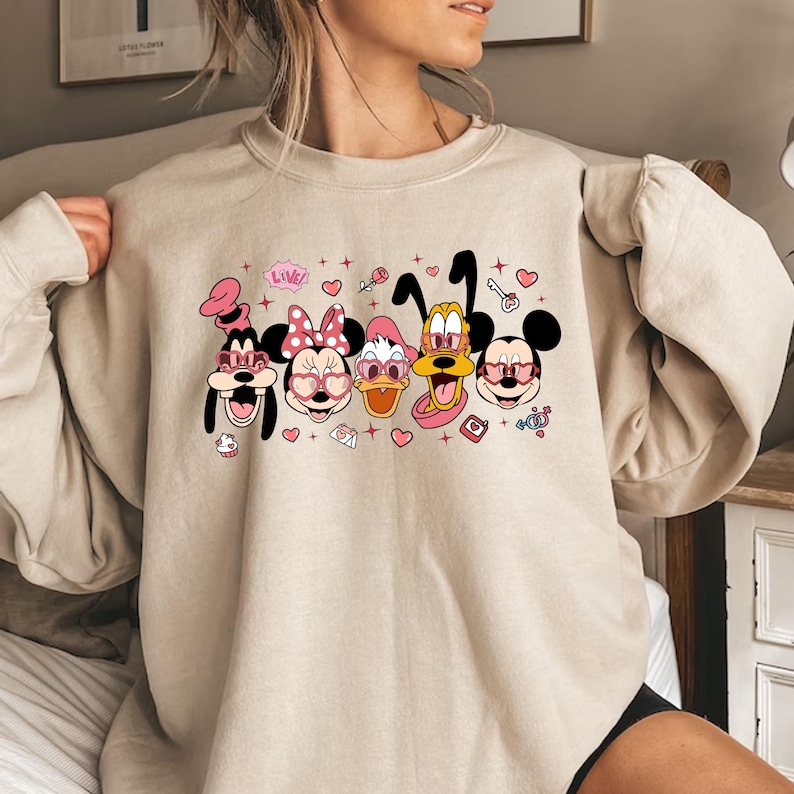 Op de afbeelding: Een beige sweatshirt met een grafische print van Mickey Mouse, Minnie Mouse, Donald Duck, Goofy en Pluto die roze hartvormige zonnebrillen dragen. De afbeelding bevat ook harten, sterren en het woord "Love!" in roze.