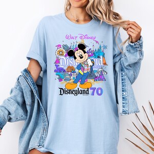 May include: Light blue t-shirt with a colorful graphic of Mickey Mouse and Disneyland attractions. The text "Walt Disney" and "Disneyland Resort 70" is printed on the shirt.