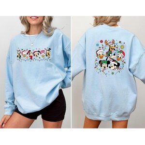 May include: Light blue sweatshirt featuring a festive design with Disney characters wearing Santa hats and Christmas lights. The front of the sweatshirt displays a line of characters, while the back showcases a circular arrangement of characters.