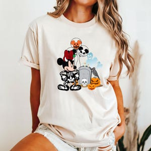 May include: Beige t-shirt featuring a Halloween-themed Minnie Mouse design. Minnie is dressed as a skeleton, holding a jack-o'-lantern, with balloons, pumpkins, a tombstone, and a crow. The design has a cartoon style.