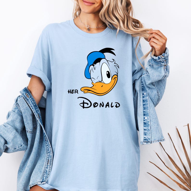 May include: Light blue t-shirt featuring a cartoon image of Donald Duck with a blue hat and orange beak. The text "HER DONALD" is printed below the image in black. The shirt has rolled-up sleeves.