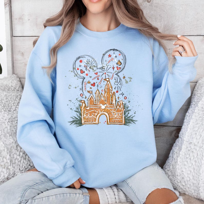 K&ouml;nnte beinhalten: Hellblaues Sweatshirt mit einem Lebkuchenschloss-Design, Mickey Mouse Ohren, Feuerwerk und festlichen Akzenten. Die Grafik ist in Braun-, Rot- und Wei&szlig;t&ouml;nen gehalten. Das Sweatshirt hat lange &Auml;rmel und einen Rundhalsausschnitt.