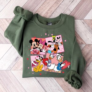 May include: Green sweatshirt with a Valentine's Day theme featuring Mickey Mouse, Minnie Mouse, Goofy, Pluto, and Donald Duck. The characters are holding hearts and surrounded by pink hearts.