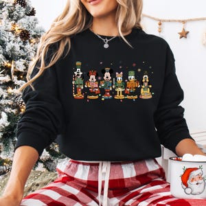 May include: Black sweatshirt featuring a colorful graphic of Disney characters as nutcrackers. The characters include Mickey Mouse, Minnie Mouse, and Donald Duck, each in festive outfits. The design is set against a backdrop of small, colorful dots.