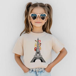 May include: A beige t-shirt featuring a black and white Eiffel Tower design with cartoon princess characters. The girl is wearing heart-shaped sunglasses with a city view.