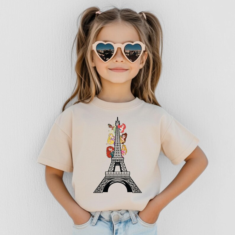 May include: A beige t-shirt featuring a black and white Eiffel Tower design with cartoon princess characters. The girl is wearing heart-shaped sunglasses with a city view.