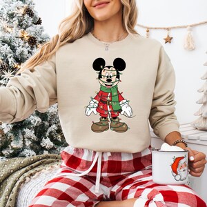 May include: Beige sweatshirt featuring a festive Mickey Mouse design. Mickey is dressed in a red outfit with a green scarf and is adorned with Christmas lights. The image also shows red and white plaid pajama pants and a mug with a Santa Claus graphic.