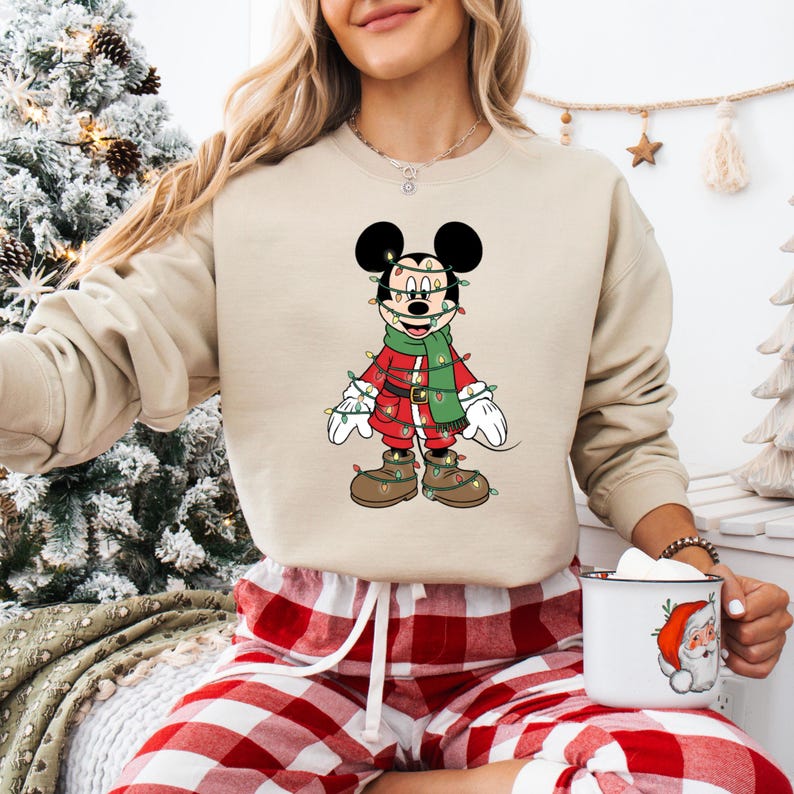 May include: Beige sweatshirt featuring a festive Mickey Mouse design. Mickey is dressed in a red outfit with a green scarf and is adorned with Christmas lights. The image also shows red and white plaid pajama pants and a mug with a Santa Claus graphic.