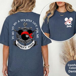 May include: Denim blue t-shirt with a pirate-themed graphic. The design features a Mickey Mouse head wearing a red bandana and a pirate hat, along with the text "Yo Ho Ho A Pirates Life For Me Disney 2026". A smaller version of the design is on the front.