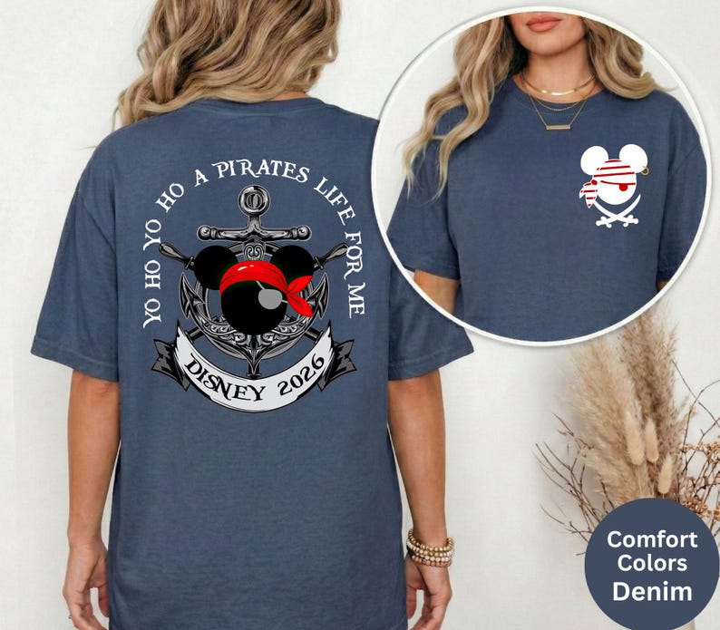 May include: Denim blue t-shirt with a pirate-themed graphic. The design features a Mickey Mouse head wearing a red bandana and a pirate hat, along with the text "Yo Ho Ho A Pirates Life For Me Disney 2026". A smaller version of the design is on the front.