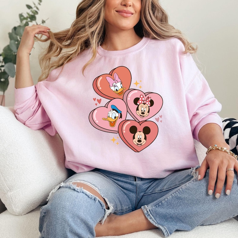 Puede incluir: Una sudadera rosa con un gr&aacute;fico de Mickey Mouse, Minnie Mouse, Donald Duck y Daisy Duck en forma de caramelo con forma de coraz&oacute;n. Los personajes sonr&iacute;en y visten sus atuendos caracter&iacute;sticos.