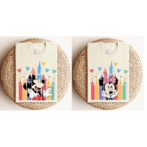 May include: Two cream colored t-shirts with a colorful castle silhouette and Mickey Mouse and Minnie Mouse cartoon characters. The castle silhouette has rainbow colored towers and pink, yellow, and blue balloons.