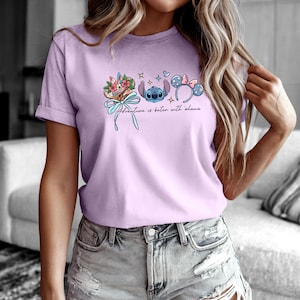 May include: A lilac t-shirt with a graphic of Stitch, floral elements, and Mickey Mouse ears. The text "Adventure is better with Ohana" is printed below the graphics. The shirt has short sleeves and a rolled cuff.