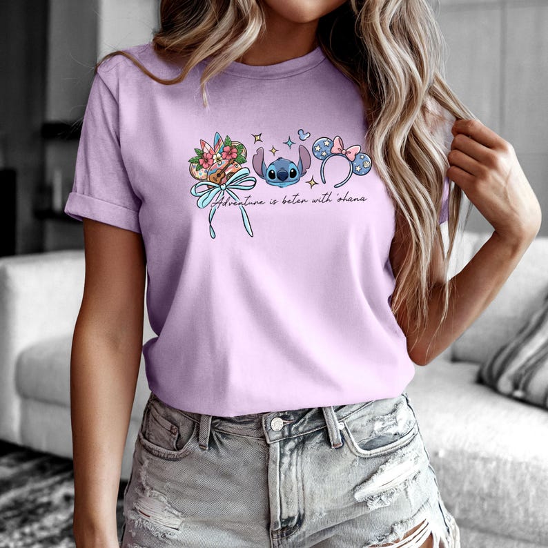 May include: A lilac t-shirt with a graphic of Stitch, floral elements, and Mickey Mouse ears. The text "Adventure is better with Ohana" is printed below the graphics. The shirt has short sleeves and a rolled cuff.