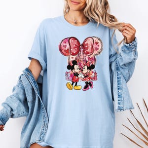 May include: Light blue t-shirt featuring a graphic of Mickey and Minnie Mouse in front of a castle. The design includes a large bow with heart accents and a pink castle. Mickey Mouse is wearing red shorts and yellow shoes, while Minnie Mouse is in a red polka dot dress.