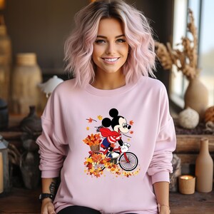 May include: A light pink sweatshirt featuring a graphic of Mickey Mouse riding a red bicycle through autumn leaves. Mickey wears a red and white scarf and a blue jacket. The sweatshirt has long sleeves and a crew neck.
