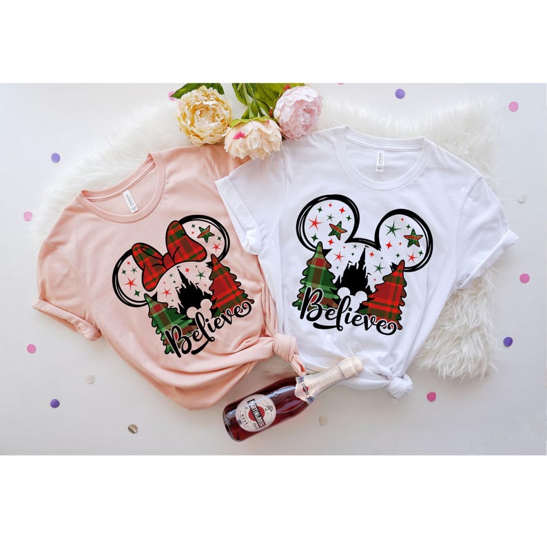 Puede incluir: Dos camisetas con un dise&ntilde;o de Mickey Mouse con tem&aacute;tica navide&ntilde;a. El dise&ntilde;o presenta las orejas de Mickey Mouse con un patr&oacute;n de cuadros rojos y verdes, un &aacute;rbol de Navidad y la palabra "Believe". Una camiseta es rosa y la otra es blanca.