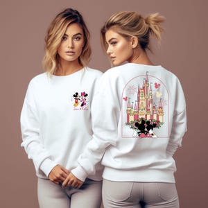 K&ouml;nnte beinhalten: Wei&szlig;er Pullover mit einer rosa und gelben Illustration von Schloss Aschenputtel mit Feuerwerk und Mickey und Minnie Mouse davor. Die Vorderseite des Pullovers zeigt eine kleinere Illustration von Mickey und Minnie Mouse mit dem Text "Jack & Emily".