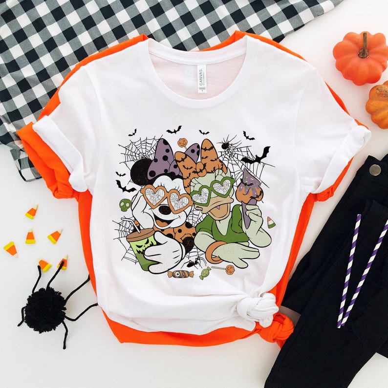 May include: White t-shirt with a Halloween-themed graphic of Mickey Mouse and Minnie Mouse wearing heart-shaped sunglasses. The graphic includes bats, spiderwebs, and candy.