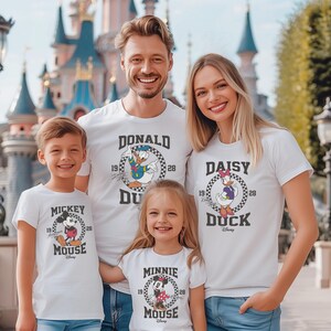 May include: A family of four wearing white t-shirts with Disney character graphics. The shirts feature Mickey Mouse, Minnie Mouse, Donald Duck, and Daisy Duck. The text on the shirts reads "Mickey Mouse", "Minnie Mouse", "Donald Duck", and "Daisy Duck".