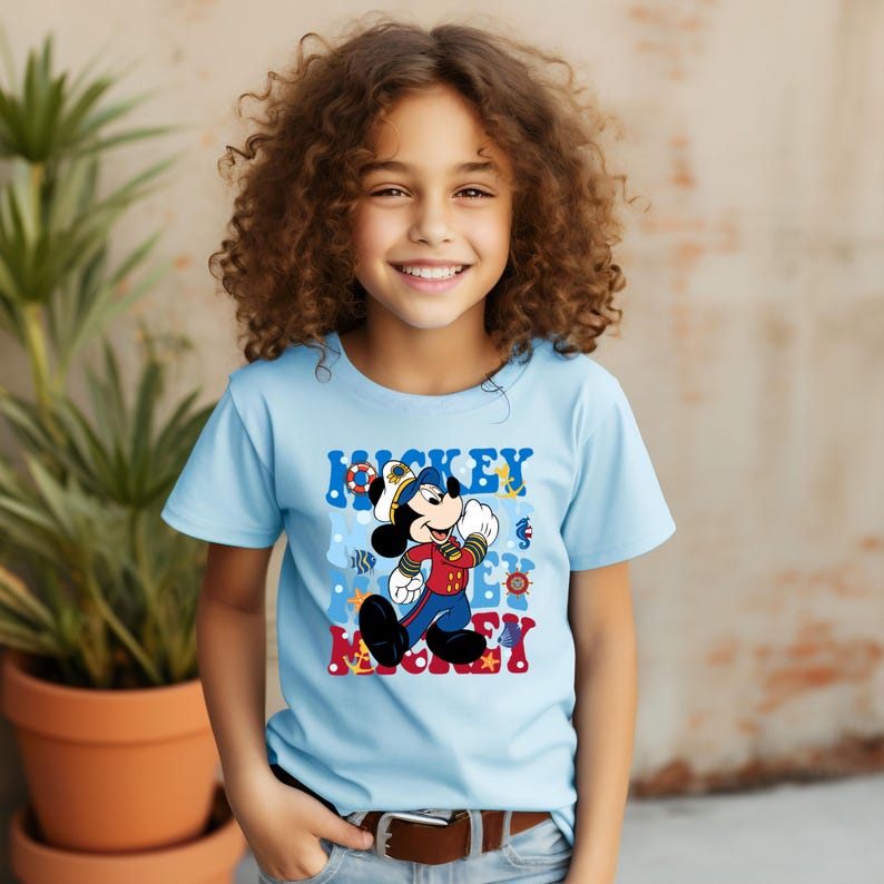 May include: A light blue t-shirt with a colorful graphic of Mickey Mouse dressed as a sailor. The graphic includes the text "Mickey Mickey" and nautical imagery like anchors, stars, and fish.