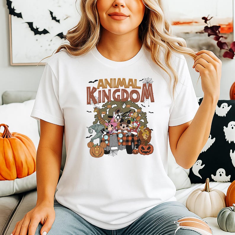 Op de afbeelding: Wit T-shirt met een kleurrijke afbeelding van stripfiguren in een vrachtwagen met de tekst "ANIMAL KINGDOM". Het ontwerp bevat pompoenen en vleermuizen, wat wijst op een Halloween-thema. Het shirt is een casual kledingstuk.