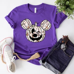 Op de afbeelding: Paars T-shirt met een wit Mickey Mouse pompoenontwerp. De pompoen is gemaakt van verschillende Halloween-themapatronen.