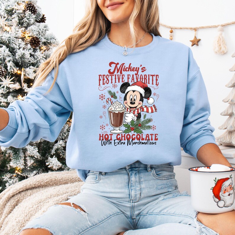 May include: Light blue sweatshirt featuring a graphic of Mickey Mouse in a Santa hat, scarf, and holding a cup of hot chocolate. The text reads "Mickey's Festive Favorite Hot Chocolate With Extra Marshmallows". The sweatshirt is a festive holiday item.