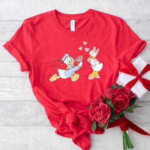 Op de afbeelding: Rood T-shirt met een afbeelding van Donald Duck die met bloemen naar Daisy Duck rent. De afbeelding is in cartoon-stijl. Het T-shirt ligt op een wit oppervlak met een bos rode rozen.