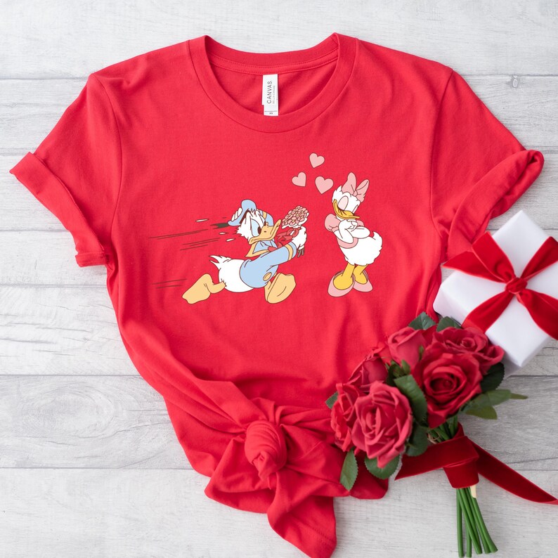 Op de afbeelding: Rood T-shirt met een afbeelding van Donald Duck die met bloemen naar Daisy Duck rent. De afbeelding is in cartoon-stijl. Het T-shirt ligt op een wit oppervlak met een bos rode rozen.
