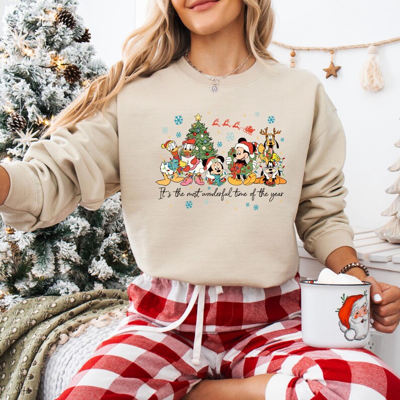 May include: A beige sweatshirt featuring a colorful Christmas graphic. The graphic includes Disney characters like Mickey and Minnie Mouse, Donald Duck, and Goofy, gathered around a Christmas tree. The phrase 'It's the most wonderful time of the year' is written below the characters. The person is wearing red and white plaid pajama pants.