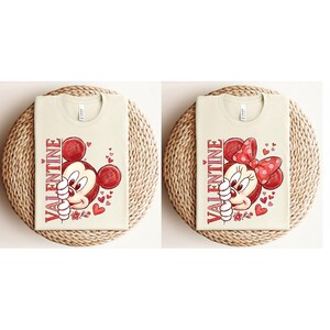 May include: Two cream-coloured t-shirts with Valentine's Day graphics. One features Mickey Mouse, the other Minnie Mouse, both with red accents and the word "VALENTINE" vertically. The shirts are on woven mats.