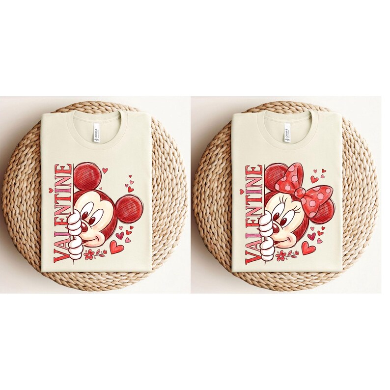 May include: Two cream-coloured t-shirts with Valentine's Day graphics. One features Mickey Mouse, the other Minnie Mouse, both with red accents and the word "VALENTINE" vertically. The shirts are on woven mats.